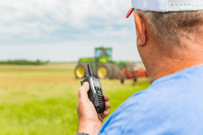 farmer holds black and silver walkie talkie while standing in field looking at tractor in distance