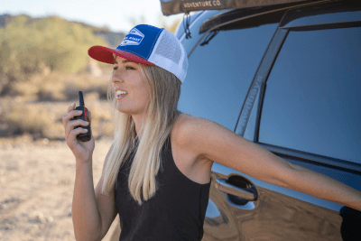 blonde woman stands by overlanding vehicle in hat while holding walkie talkie