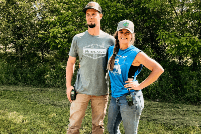 couple stands in front of trees on farm with walkie talkies attached to hip