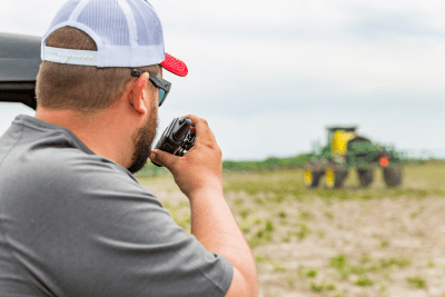 man uses walkie talkie in field while look at tractor in distance