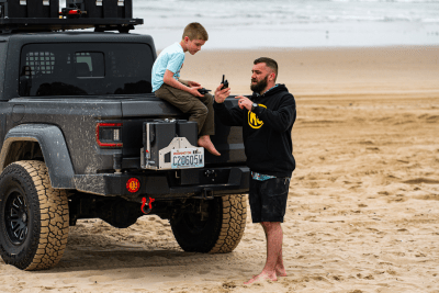 Boy sits on tailgate of Jeep with his dad and walkie talkie on a beach
