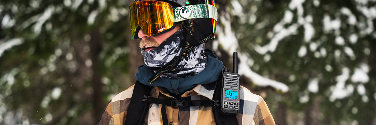 Person wearing goggles and a plaid jacket with a walkie-talkie in a snowy forest setting